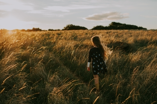 Young fashionable girl walking through long grass in paddock at sunset - Australian Stock Image