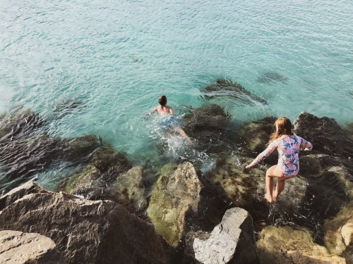 Young girl and boy climbing on coastal rocks and jumping into ocean