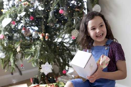 Young girl happy and smiling after opening gift in front of Christmas tree - Australian Stock Image