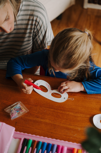 Young girl in superhero costume colouring her mask with mum sitting beside her. - Australian Stock Image