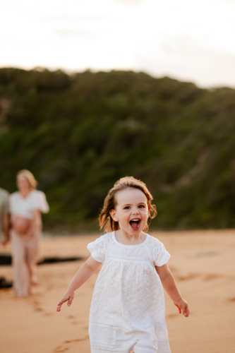 Young girl in white dress running on sandy beach at sunset while family walks behind her - Australian Stock Image