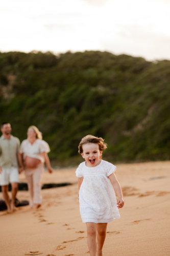 Young girl in white dress runs excitedly on sandy beach while family walks behind her at sunset - Australian Stock Image