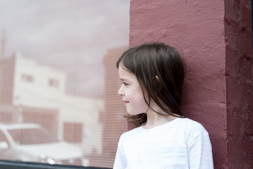 Young girl looking away, leaning against a pink brick wall with a window reflecting the street - Australian Stock Image
