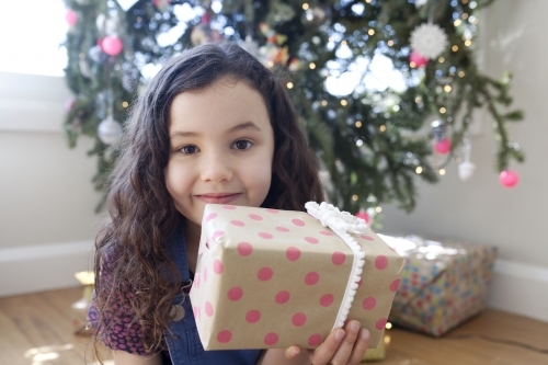 Young girl sitting in front of christmas tree holding a present up in front of her face