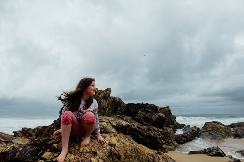 Young girl sitting on rocks at beach, wind blowing her hair