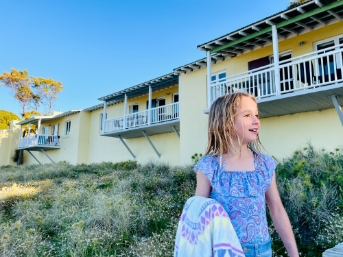 Young girl smiling in front of coastal holiday units in swimsuit holding towel - Australian Stock Image