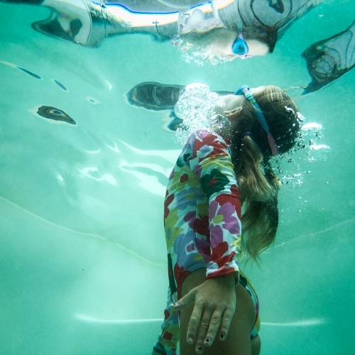 Young girl swimming to the surface underwater in pool wearing floral swimming suit and goggles - Australian Stock Image