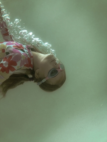 Young girl swimming underwater in pool wearing floral swim suit and goggles blowing bubbles - Australian Stock Image