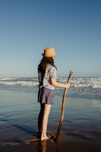 Young girl wearing a sunhat holding a stick on a beach in the afternoon - Australian Stock Image