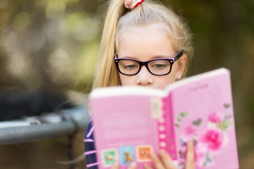 Young girl with glasses reading a book - Australian Stock Image