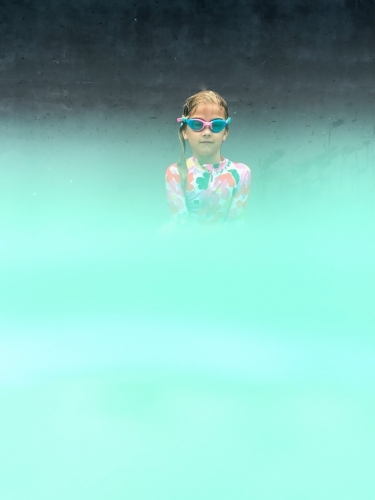 Young girls upper torso in swimming outfit and goggles on edge of pool and water in foreground - Australian Stock Image