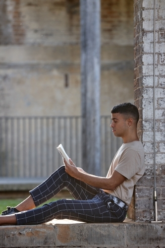 Young Indigenous man sitting against brick wall reading book - Australian Stock Image