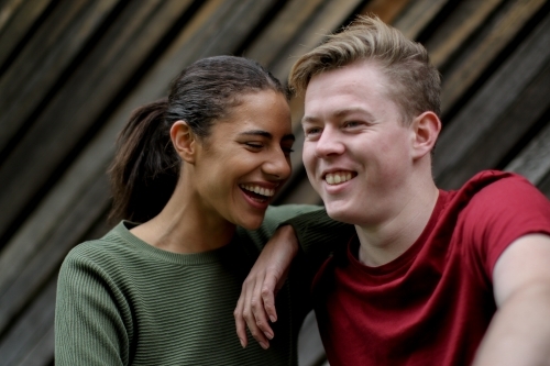 Young interracial couple laughing together in front of textured wooden panels - Australian Stock Image