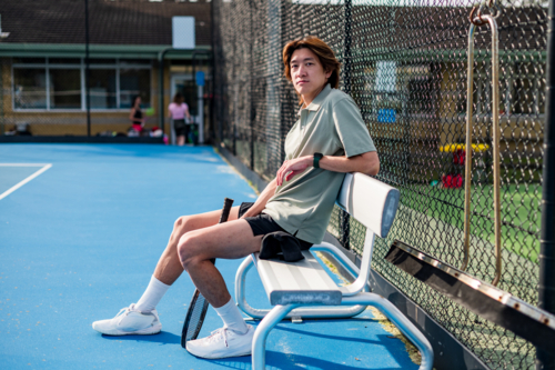 Young male athlete sits on bench beside tennis court, preparing for practice with racquet nearby - Australian Stock Image