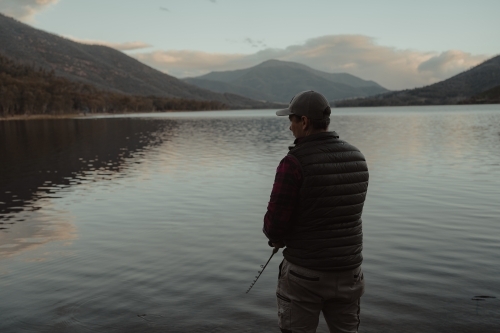 Young man fishing next to a calm lake with mountains in the background - Australian Stock Image
