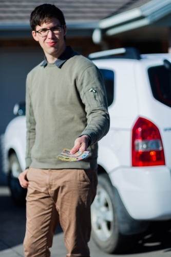 Young man holding cash standing in front of car paying up front - Australian Stock Image