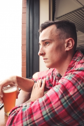 Young man holding craft beer staring out of window from bar