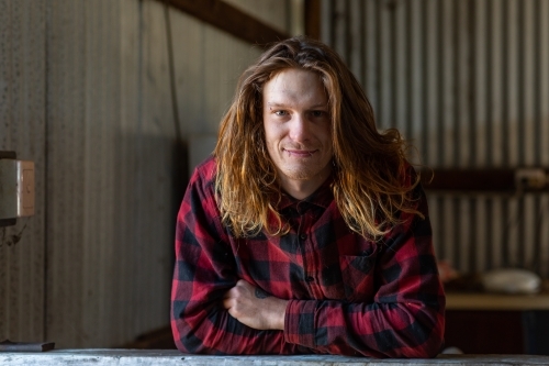 young man in flannel shirt leaning on bench in shed - Australian Stock Image