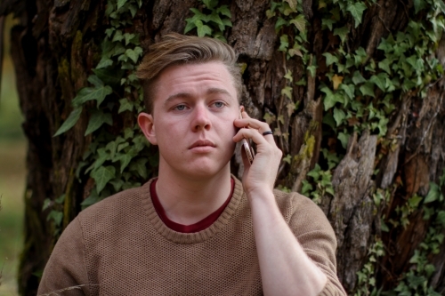 Young man on the phone sitting against a tree with a sad expression - Australian Stock Image