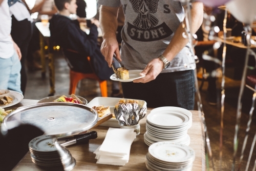 Young man putting food from a serving table on a plate, people in background at a party - Australian Stock Image