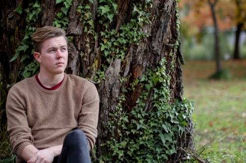 Young man sitting outdoors at the base of a tree contemplating life - Australian Stock Image