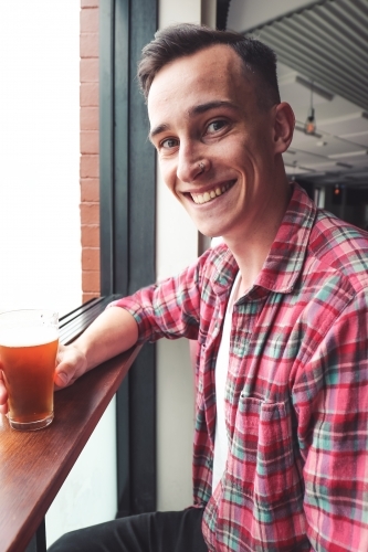 Young man smiling at a window ledge of a bar holding a craft beer