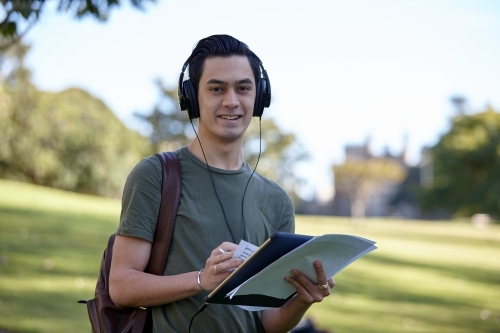 Young man with dark hair outside holding device wearing headphones at park - Australian Stock Image