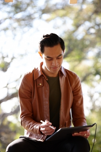 Young man with dark hair outside writing on device at park - Australian Stock Image