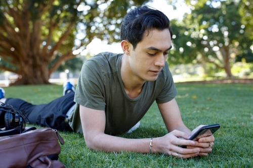 Young man with dark hair using mobile phone on grass at park