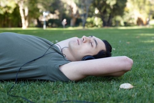 Young man with eyes closed relaxing listening to headphones at park - Australian Stock Image