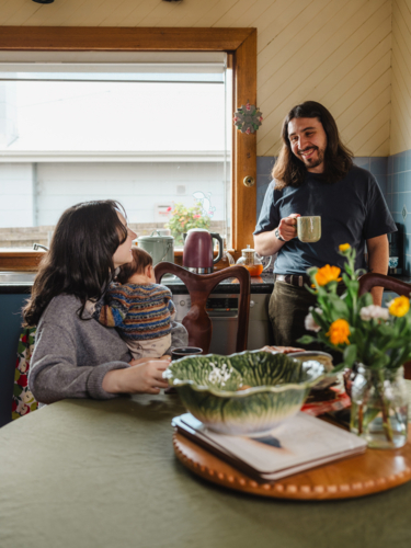 Young mother holding baby while seated and father standing drinking coffee - Australian Stock Image