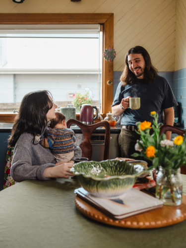Young mother holding baby while seated and father standing drinking coffee in kitchen at home - Australian Stock Image