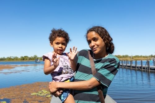 young mother holding daughter on her hip with water in background - Australian Stock Image