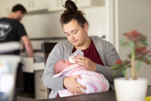 Young mum bottle-feeding baby with father in kitchen - Australian Stock Image