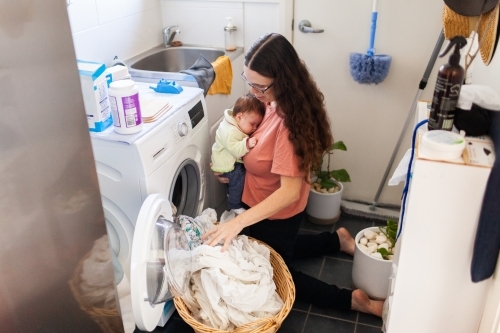 Young mum taking wet clean clothes out of washing machine while holding newborn baby - Australian Stock Image