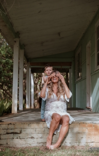 Young son covering his mother's eyes and playing with her on the front porch of home - Australian Stock Image