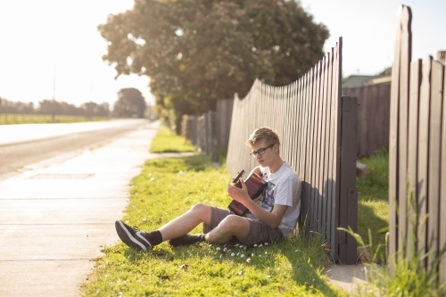 Young teen boy playing acoustic guitar by fence - Australian Stock Image