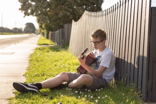 Young teen boy playing acoustic guitar by fence - Australian Stock Image