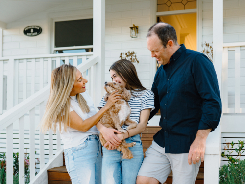 Young teenage girl carrying a dog while standing beside her parents outside the house. - Australian Stock Image