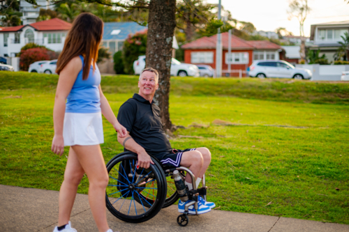 Young woman approaches smiling man in wheelchair, sharing a moment in afternoon sunlight at park - Australian Stock Image