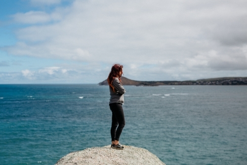 Young woman arms crossed standing on rock looking at ocean - Australian Stock Image