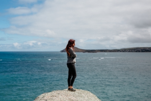 Young woman arms crossed standing on rock looking at ocean