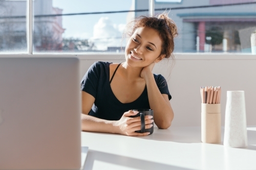 Young woman having coffee at a desk smiling at her computer screen