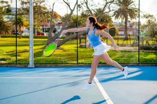Young woman hits forehand shot on tennis court surrounded trees during daylight - Australian Stock Image