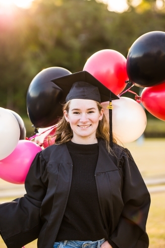 Young woman holding balloons wearing graduation cap and gown smiling outside - Australian Stock Image