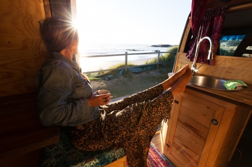 Young woman in camper van watching summer sunrise while drinking coffee by the beach
