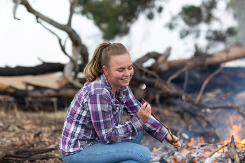 Young woman in checked shirt and jeans toasting marshmallows on campfire