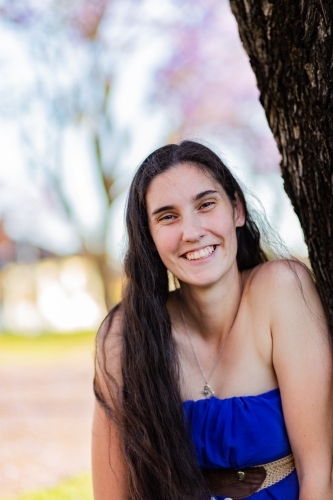 Young woman in her twenties smiling outside leaning on tree - Australian Stock Image
