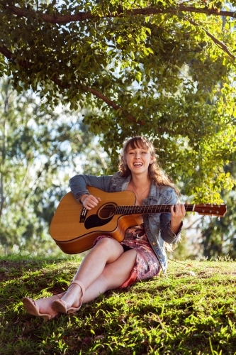 Young woman on grassy hill playing guitar laughing - Australian Stock Image