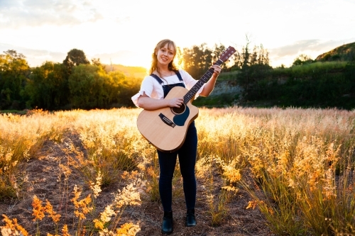 Young woman playing guitar in grass backlit by golden light - Australian Stock Image